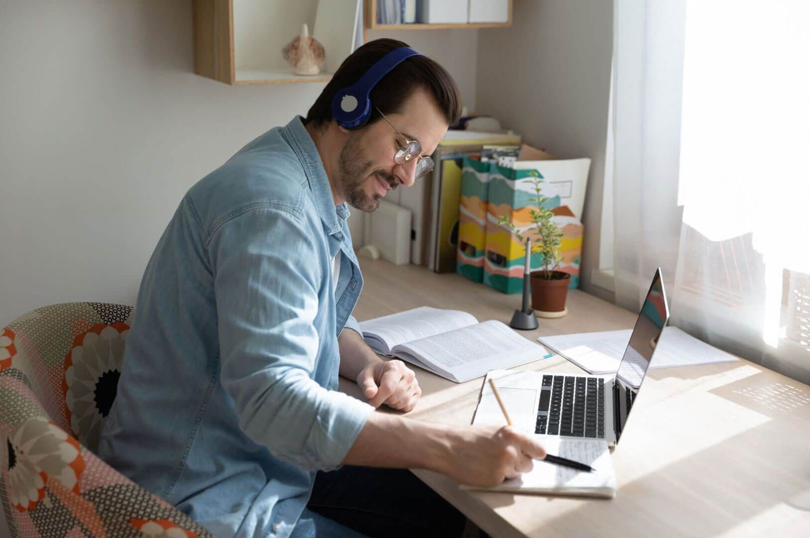 Hombre con audífonos estudiando y tomando notas frente a laptop en escritorio