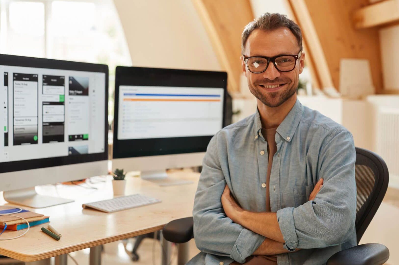 Hombre con gafas sonriendo de brazos cruzados frente a monitores con diseño de aplicaciones